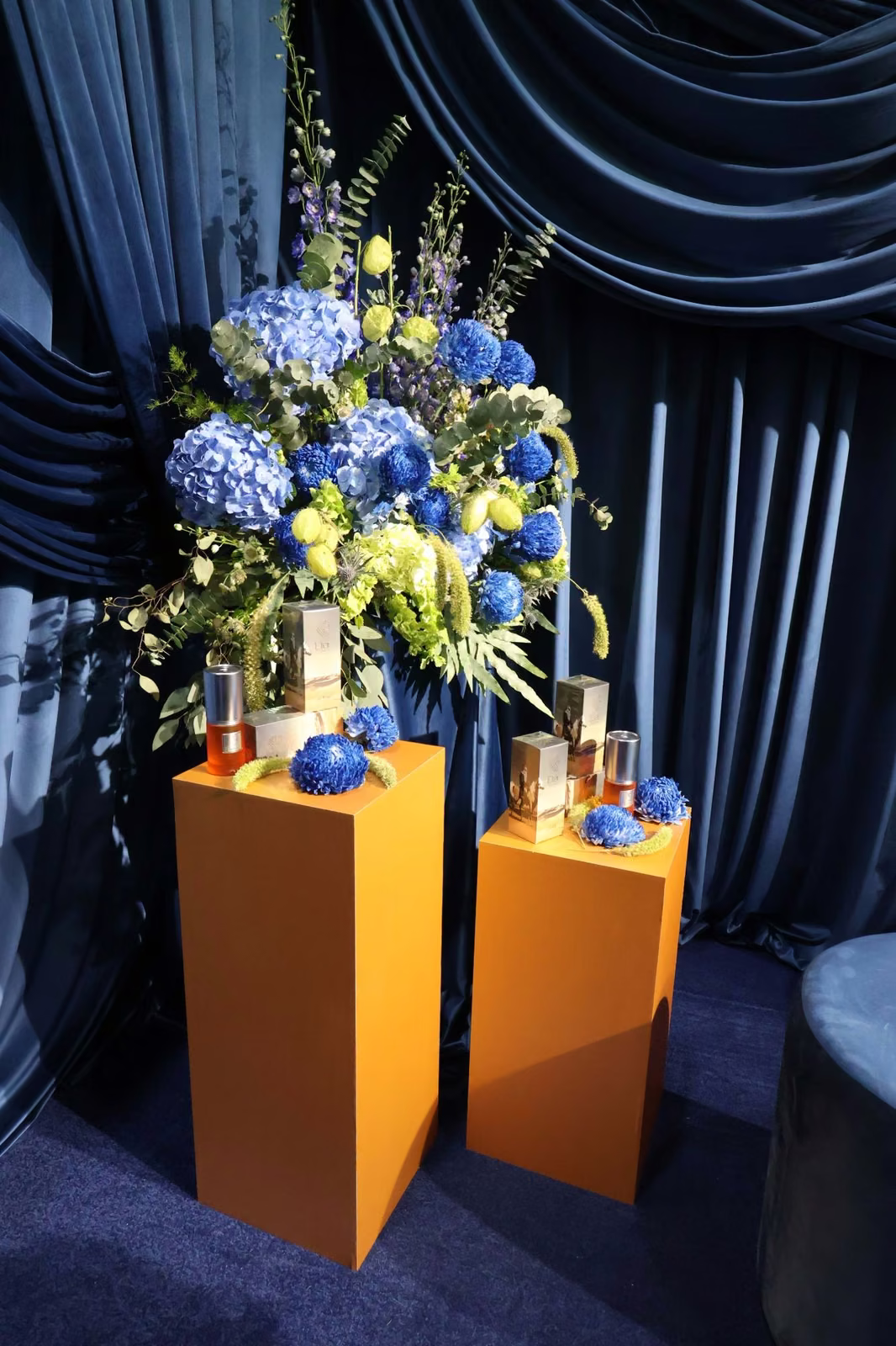 Etèr Fragrance bottles on copper-toned plinths with a blue hydrangea arrangement against navy fire-retardant draping
